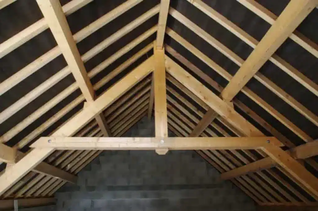 Charpente bois : construction de ferme et pose de toiture Charpente en bois neuve avec ferme et entrait, supportant des chevrons. Membrane noire de sous-toiture visible. Murs en blocs de béton gris.